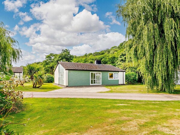A small, single-story house with a green exterior, a driveway, and a lush garden with a weeping willow tree. - 2 Bed in Saltash (oc-88308) (Notter)