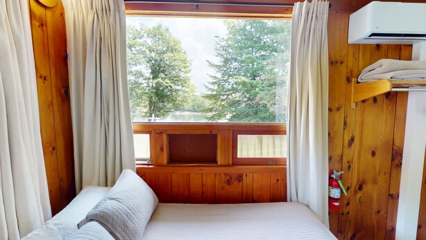 Habitación de cabaña de madera con cama, ventana con cortinas y vistas a árboles, además de un aire acondicionado instalado.