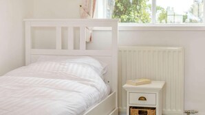 Un letto bianco con testiera, un comodino con un libro, un termosifone e una finestra con vista sul verde.