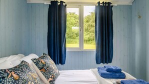 Ett rum med en säng, två fönster med gardiner, utsikt över grönområden utanför och ett litet bord med handdukar.