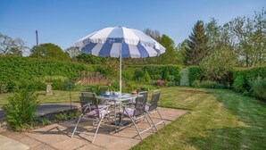 Une terrasse extérieure avec une table et des chaises, un parasol à rayures et un jardin agrémenté de diverses plantes et d’une structure en pierre.