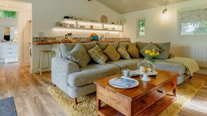 Een gezellige woonkamer met een grijze bank, een houten salontafel en een keukenhoek met witte kasten en een houten balk.
