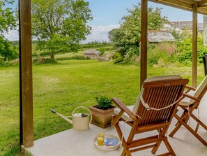A covered outdoor seating area with a wooden chair, a watering can, and a tray with a book and glasses.