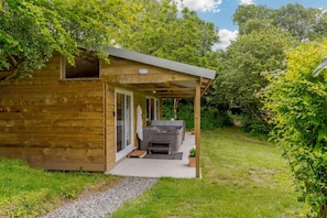 A wooden cabin with a covered patio, a hot tub, and a view of a lush garden.