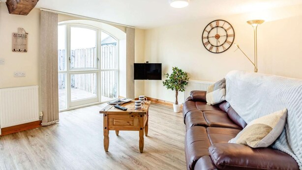 Un salon avec une table basse en bois, un canapé en cuir marron, un téléviseur fixé au mur, une horloge et une grande fenêtre.