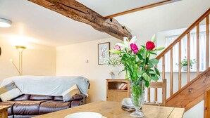 Une table à manger en bois, sur laquelle se trouvent un saladier blanc et un verre à vin, un canapé en cuir avec une couverture, et un escalier en bois.