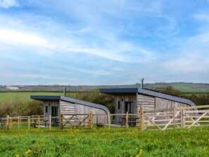 Two rustic wooden cabins with metal roofs, surrounded by a wooden fence, set in a green field with a clear sky.