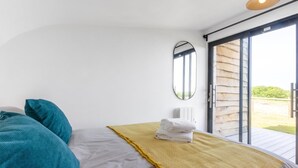 Ein Schlafzimmer mit einem Bett, einer gelben Tagesdecke, türkisfarbenen Kissen, einem Spiegel und einer Schiebetür zu einem Außenbereich.