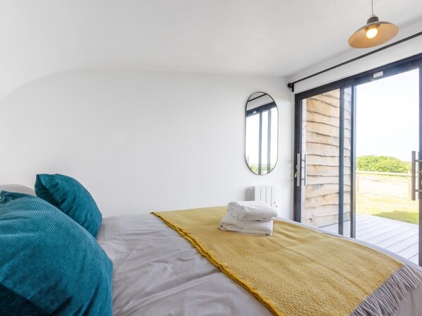 A bedroom with a bed, a yellow throw blanket, teal pillows, a mirror, and a sliding glass door to an outdoor area.