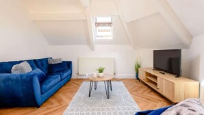 Ein Loft-Wohnzimmer mit blauem Sofa, einem Holztisch, einem Fernseher auf einem Holzständer und einem gemusterten Teppich.