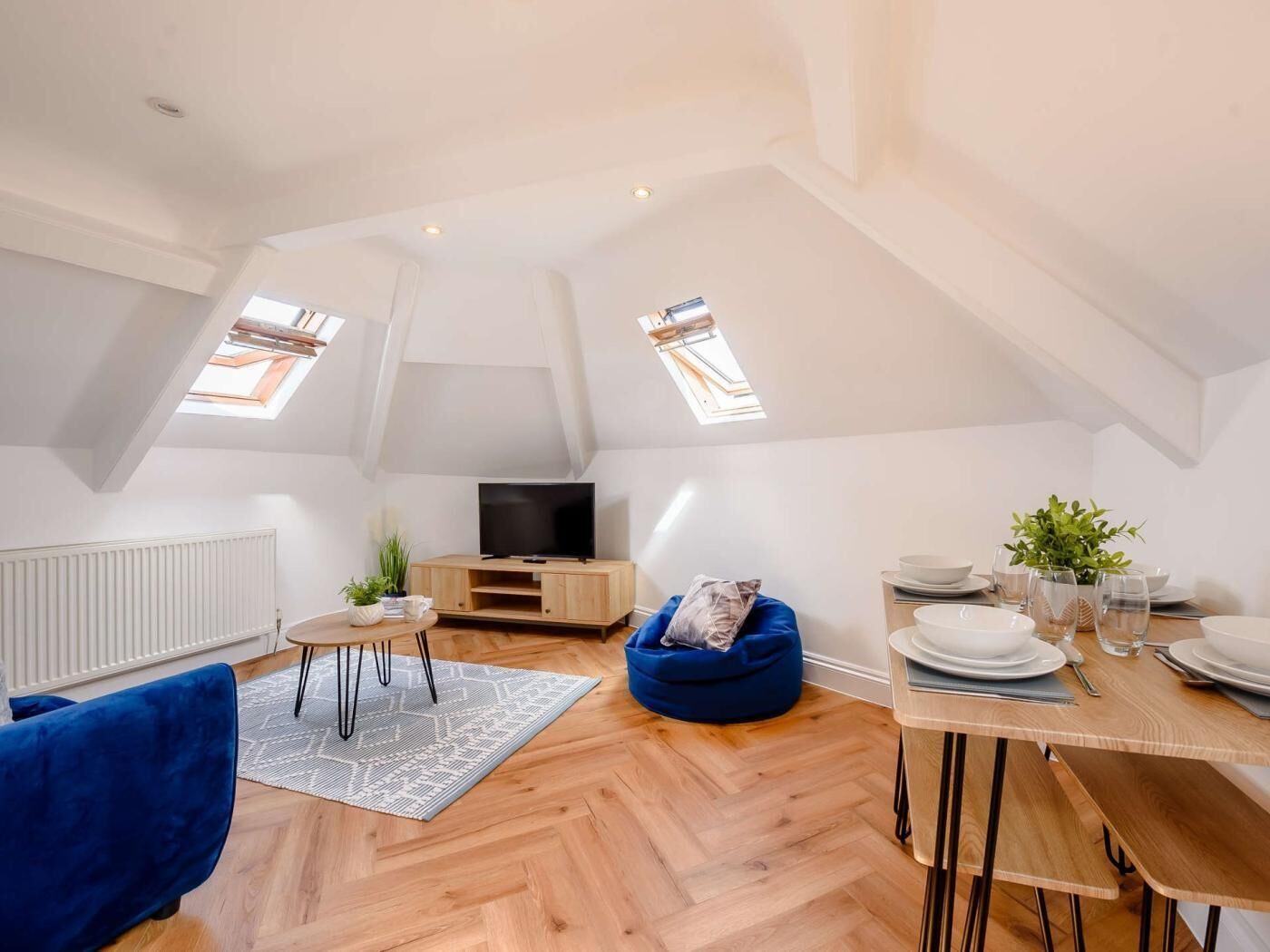 Un appartement moderne de style « loft », avec un plafond haut, un plancher en bois, un canapé bleu confortable, un coin repas avec une table dressée pour deux personnes et un téléviseur posé sur un meuble en bois.