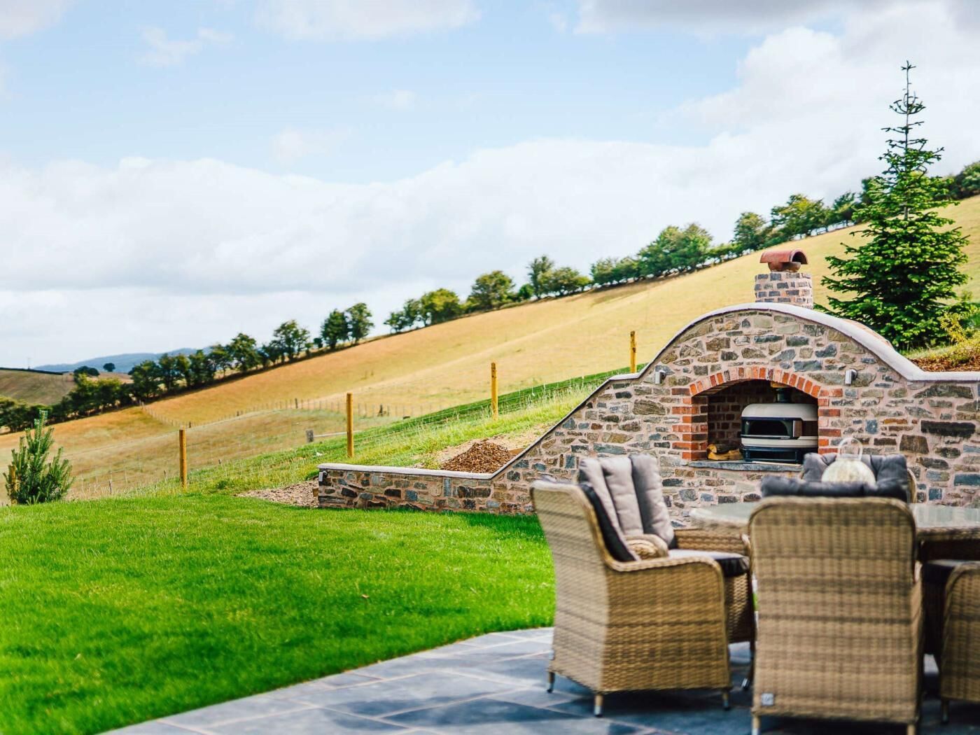 Une terrasse extérieure aménagée avec des meubles en pierre et un four en briques, le tout surplombant un paysage de collines douces et un ciel dégagé.