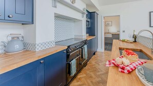 A modern kitchen with blue cabinets, a wooden countertop, a stove, and a dining area with a table set for two. - 3 Bed in Montgomery (oc-88641) (Llandyssil)