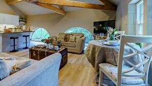 Un accogliente soggiorno mansardato con travi a vista in legno, una zona pranzo con tavolo apparecchiato per due e una cucina sullo sfondo.