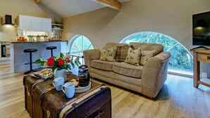 Un accogliente soggiorno mansardato con soffitto in legno, una cucina con mobili bianchi, un divano con cuscini decorati, una valigia vintage e un'ampia finestra con vista sul verde.