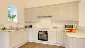 A modern kitchen with white cabinets, a built-in oven, a microwave, and a window with a flower vase. - 2 Bed in Askam-in-furness (oc-88651) (Kirkby Ireleth)