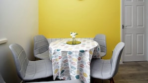 Un comedor con una mesa cubierta por un mantel estampado, cuatro sillas grises, un reloj blanco en la pared y una puerta cerrada.