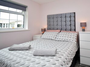 Un lit bien fait, avec une couvre-lit à motifs géométriques et des oreillers, une tête de lit capitonnée, des lampes de chevet et une fenêtre donnant sur des immeubles.