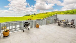 Eine Terrasse mit Blick auf grüne Felder und ein Gewässer, ein Grill und Gartenmöbel.