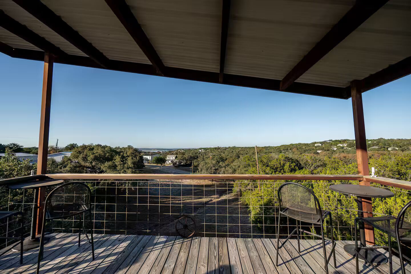 A covered outdoor area with a wooden deck, metal chairs, and a view of a landscape with trees and buildings.