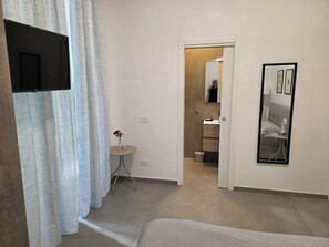Une chambre d’hôtel avec un lit, une petite table sur laquelle est posé un vase de fleurs, un miroir et une salle de bain visible à travers une porte ouverte.