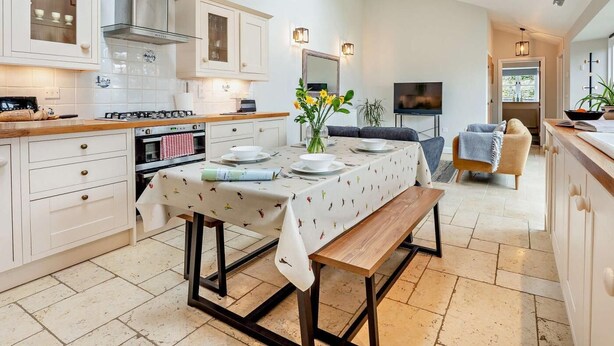 A modern kitchen with white cabinetry, a dining table with a floral tablecloth, and a TV in the living area. - 2 Bed in Ventnor (oc-88857) (Niton)