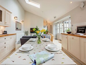 A modern kitchen with a dining area, featuring a central island with a vase of flowers, a flat-screen TV mounted on the wall, and a chalkboard sign. - 2 Bed in Ventnor (oc-88857) (Niton)