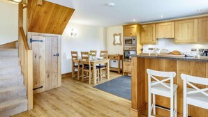 A kitchen with wooden cabinets, a dining area with chairs, a staircase, and a kitchen island. - 2 Bed in Launceston (oc-88859) (Saint Giles on the Heath)