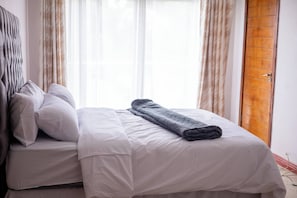 A neatly made bed with white linens and a folded blanket, a wooden door, and sheer curtains allowing natural light. - Room-luxurious Room - D (Johannesburg)