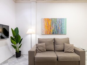Een moderne woonkamer met een beige bank, een potplant, een vloerlamp en een kleurrijk abstract schilderij aan de muur.