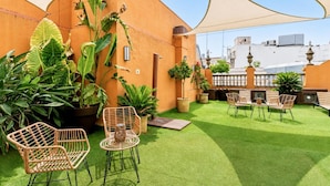 Terraza en la azotea con césped artificial, mobiliario de mimbre, plantas en macetas y una gran sombrilla que proporciona sombra.