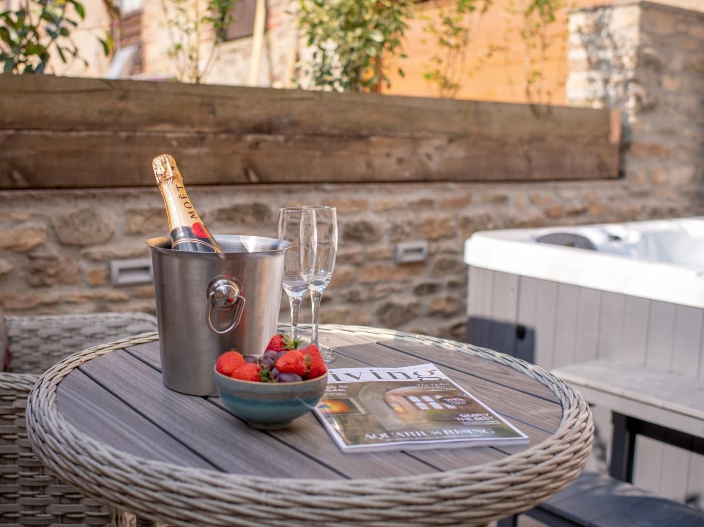A wicker table with a metal bucket containing a bottle of champagne, two empty glasses, a bowl of strawberries, and a magazine.