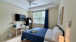 Une chambre d’hôtel comprenant un lit, un bureau, une chaise, un ventilateur de plafond, une fenêtre avec des rideaux, un téléviseur fixé au mur et une lampe de chevet.