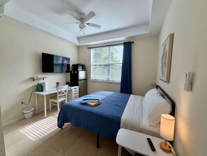 Ein Hotelzimmer mit einem Bett, einem Schreibtisch, einem Stuhl, einem Deckenventilator, einem Fenster mit Vorhängen, einem an der Wand befestigten Fernseher und einer Nachttischlampe.