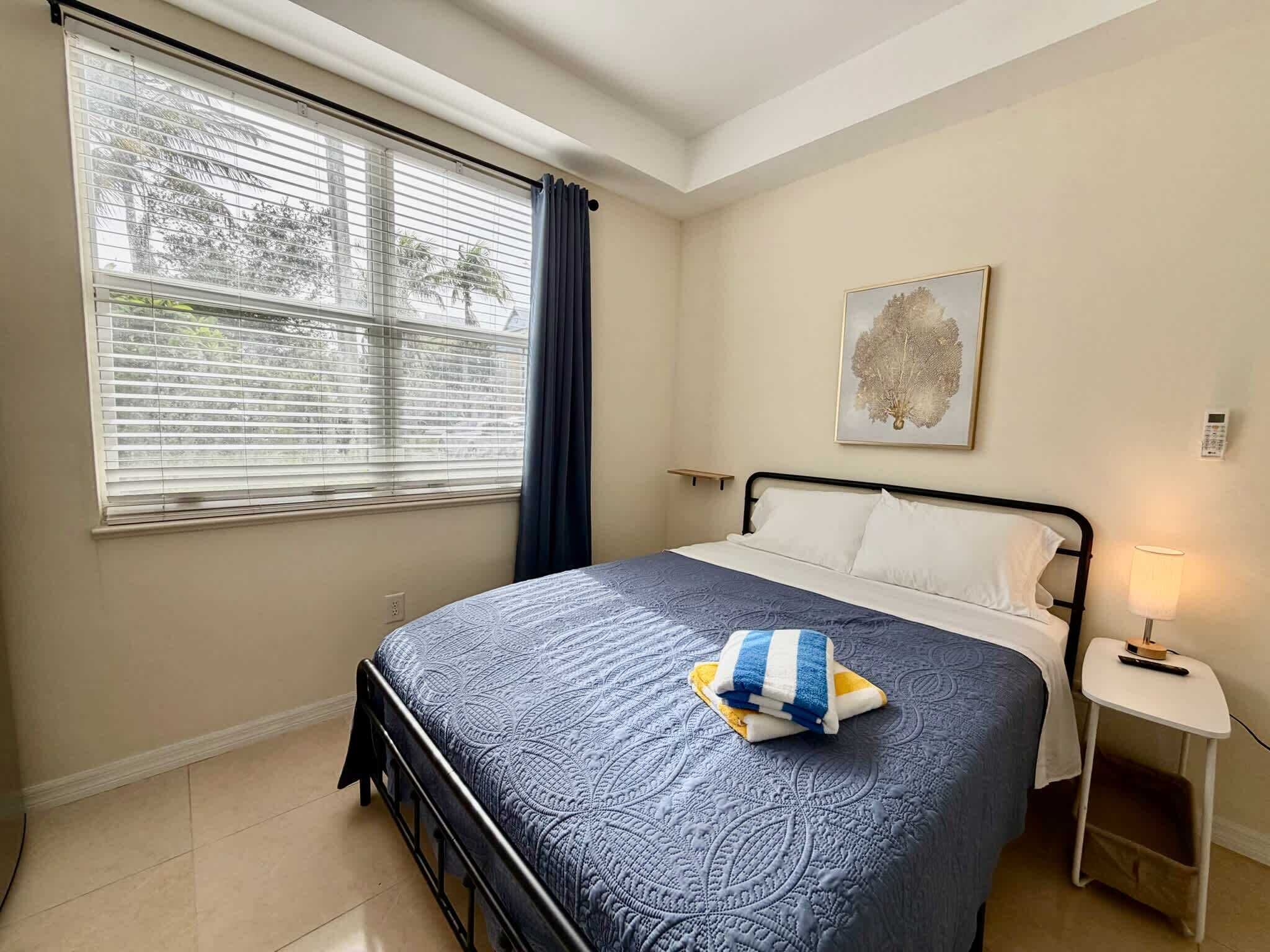 A bedroom with a bed, a nightstand, a lamp, a window with blinds, and a wall-mounted painting.