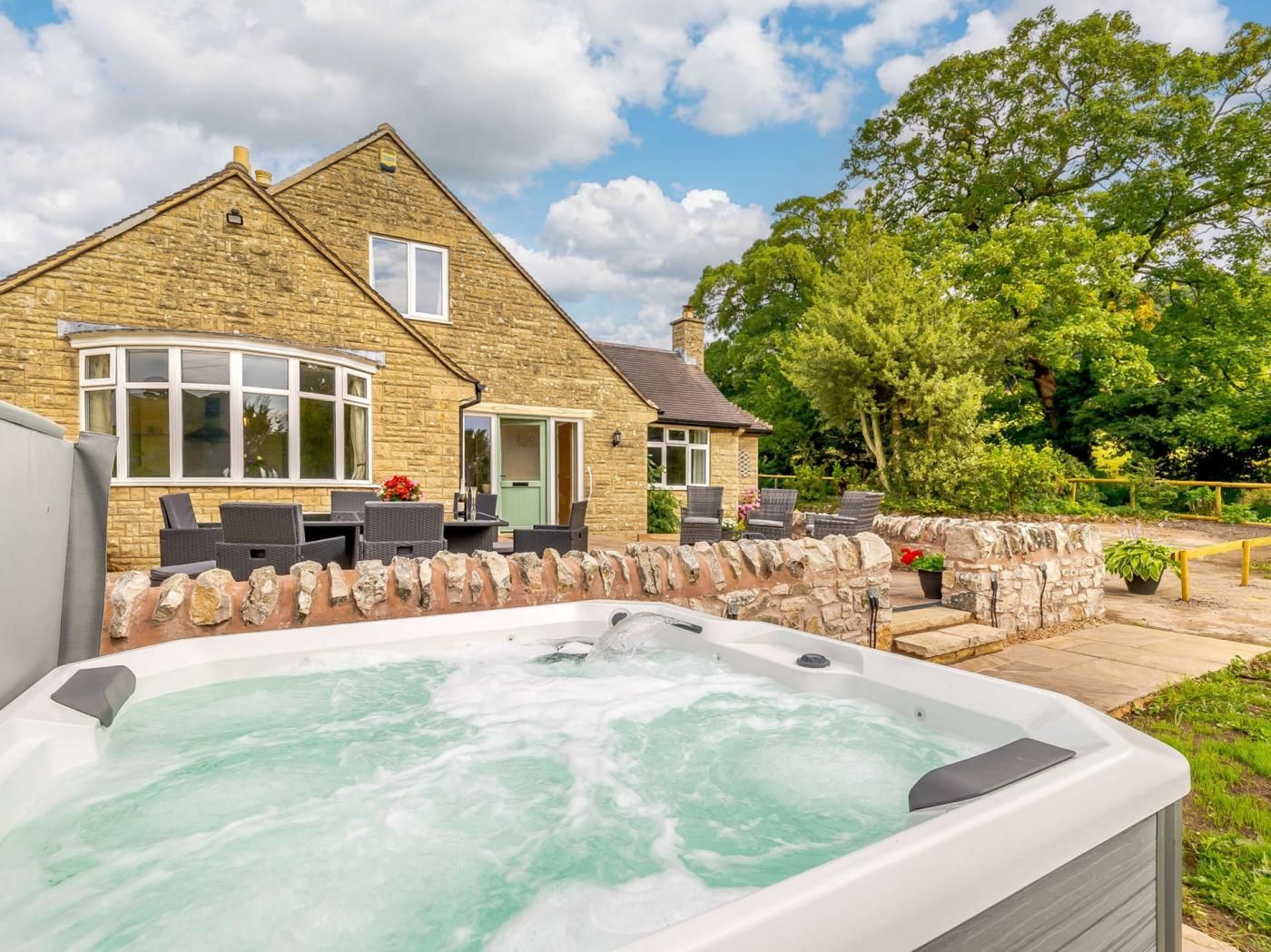 A stone house with a hot tub in the foreground, a patio with outdoor furniture, and a large tree in the background.