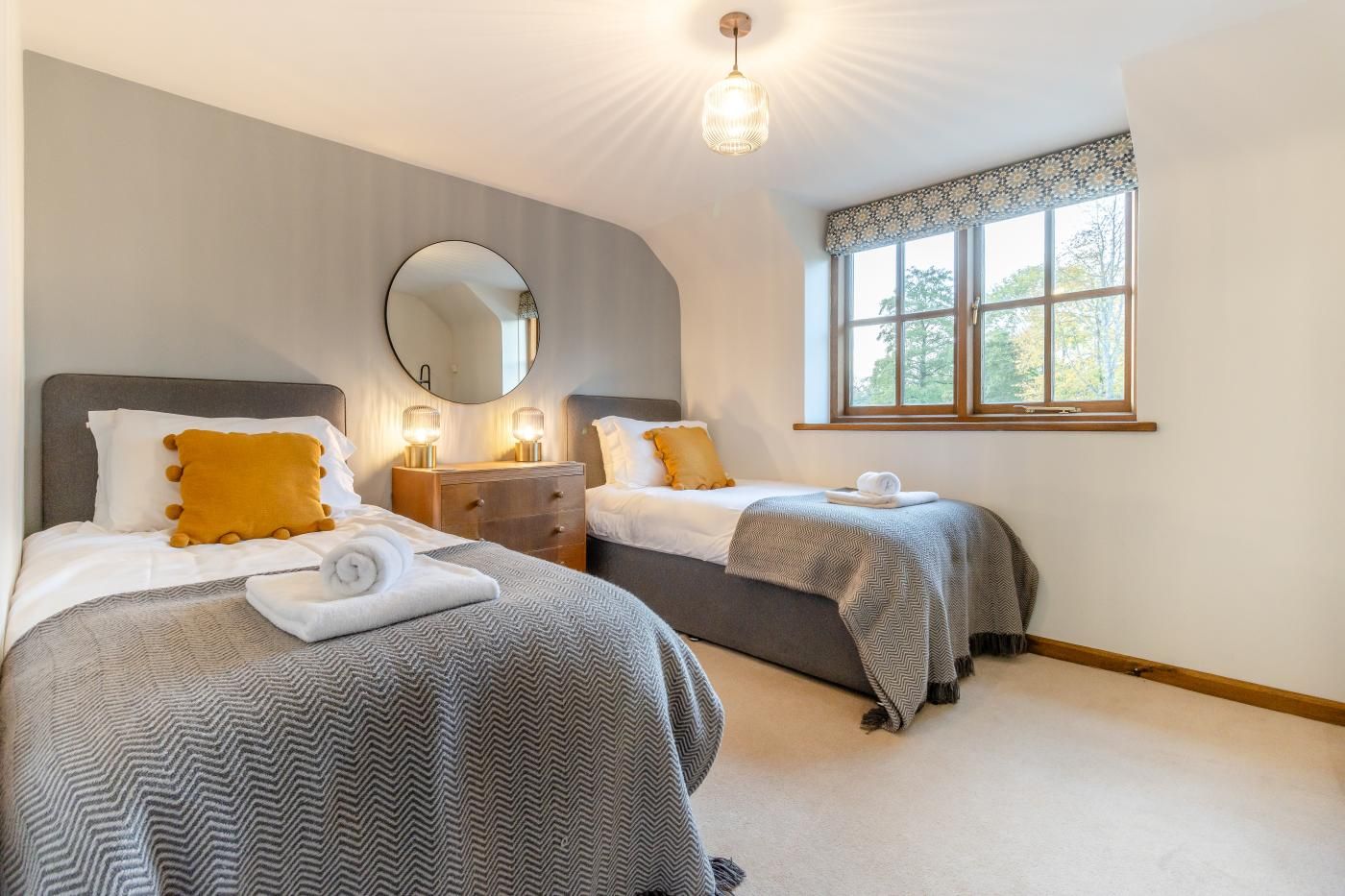 A hotel room with two beds, a round mirror, bedside lamps, a dresser, and a window with a view of trees.