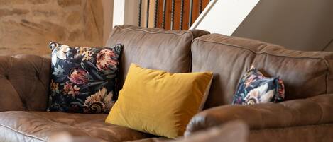 A brown leather sofa with floral and mustard-colored cushions in a room with stone walls and a wooden staircase.