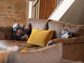A brown leather sofa with floral and mustard-colored cushions in a room with stone walls and a wooden staircase. - 1 Bed in Richmond (oc-88588) (Brompton on Swale)