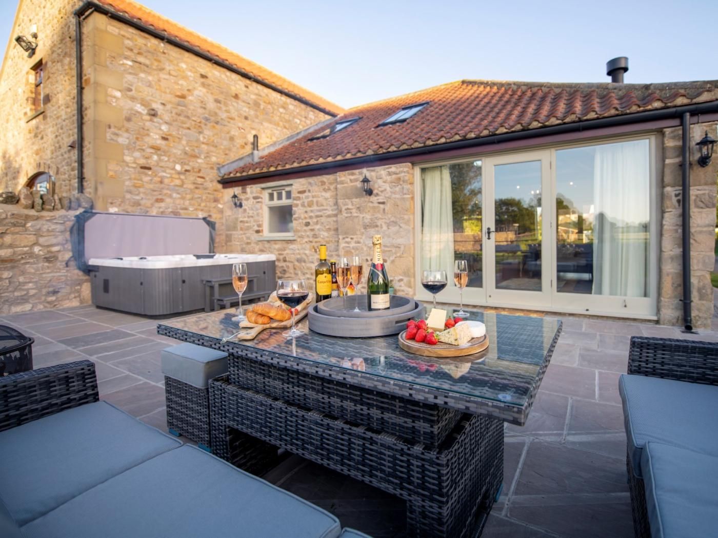 An outdoor patio with a hot tub, wicker furniture, and a table set with wine glasses, bottles, cheese, and strawberries.