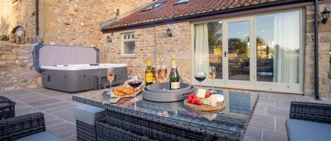An outdoor patio with a hot tub, wicker furniture, and a table set with wine glasses, bottles, cheese, and strawberries.