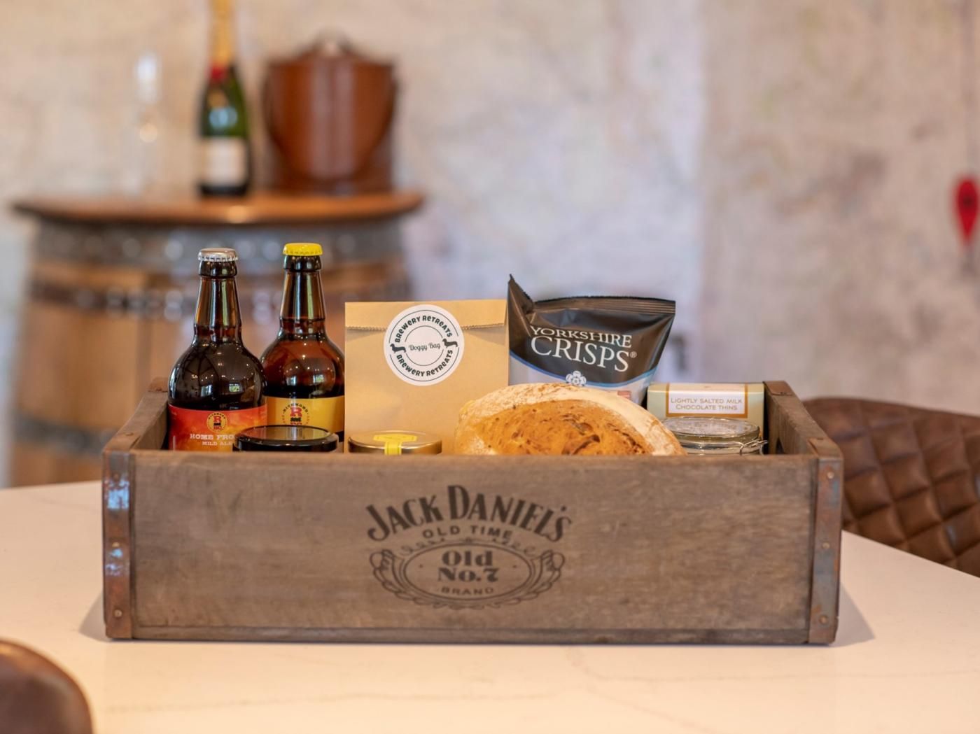 A wooden crate containing a bottle of Jack Daniel's whiskey, a bottle of beer, a bag of Yorkshire crisps, a chocolate tin, and a box of biscuits.