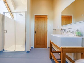 Ein Badezimmer mit Duschkabine aus Glas, einer Holztür, einem weißen Waschbecken mit Spiegel darüber und einer hölzernen Sitzbank.
