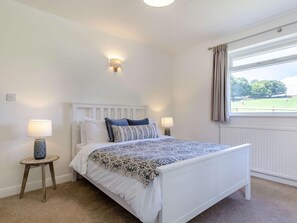 Un dormitorio con cama, dos lámparas de noche, una luz en la pared, una ventana con cortinas y vistas a un campo verde.