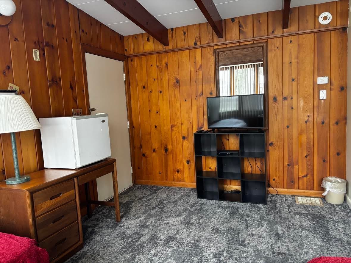 Una habitación con paneles de madera, un refrigerador blanco, un mueble negro para entretenimiento y un escritorio pequeño con cajones.