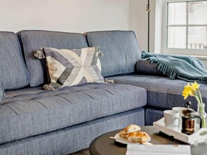 A grey sofa with a patterned pillow, a blue throw blanket, a coffee table with a tray of pastries, and a vase with yellow flowers. - 2 Bed in Braunton (oc-87750) (Croyde)