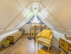 Ein kleines Zimmer im Dachgeschoss mit schrägen Wänden, einer Holzbank, einem Stuhl mit Kissen, einem kleinen Tisch und einer Stehlampe.