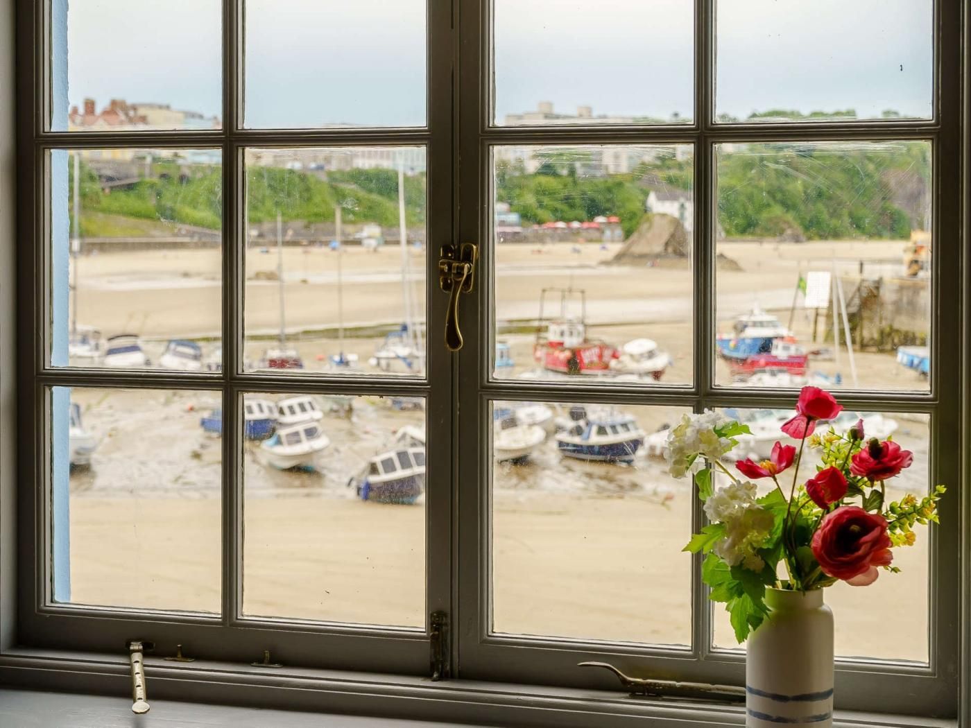 Una ventana con vistas a barcos en una orilla arenosa, un mecanismo de cierre y un florero con flores.
