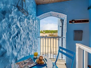 Une pièce avec un mur bleu à la texture particulière, un balcon donnant sur la plage, une table avec un journal, un verre de jus d’orange et un croissant.