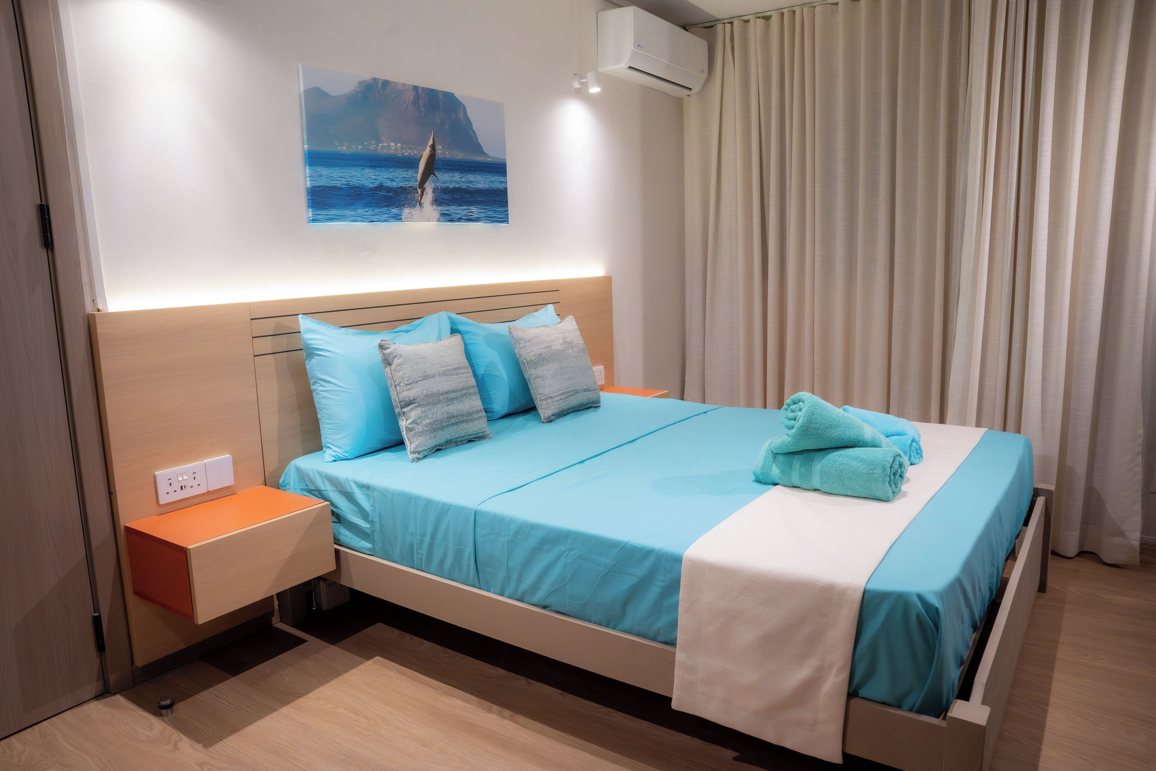 A hotel room with a bed, a nightstand, a wall-mounted air conditioner, and a painting of a coastal scene.